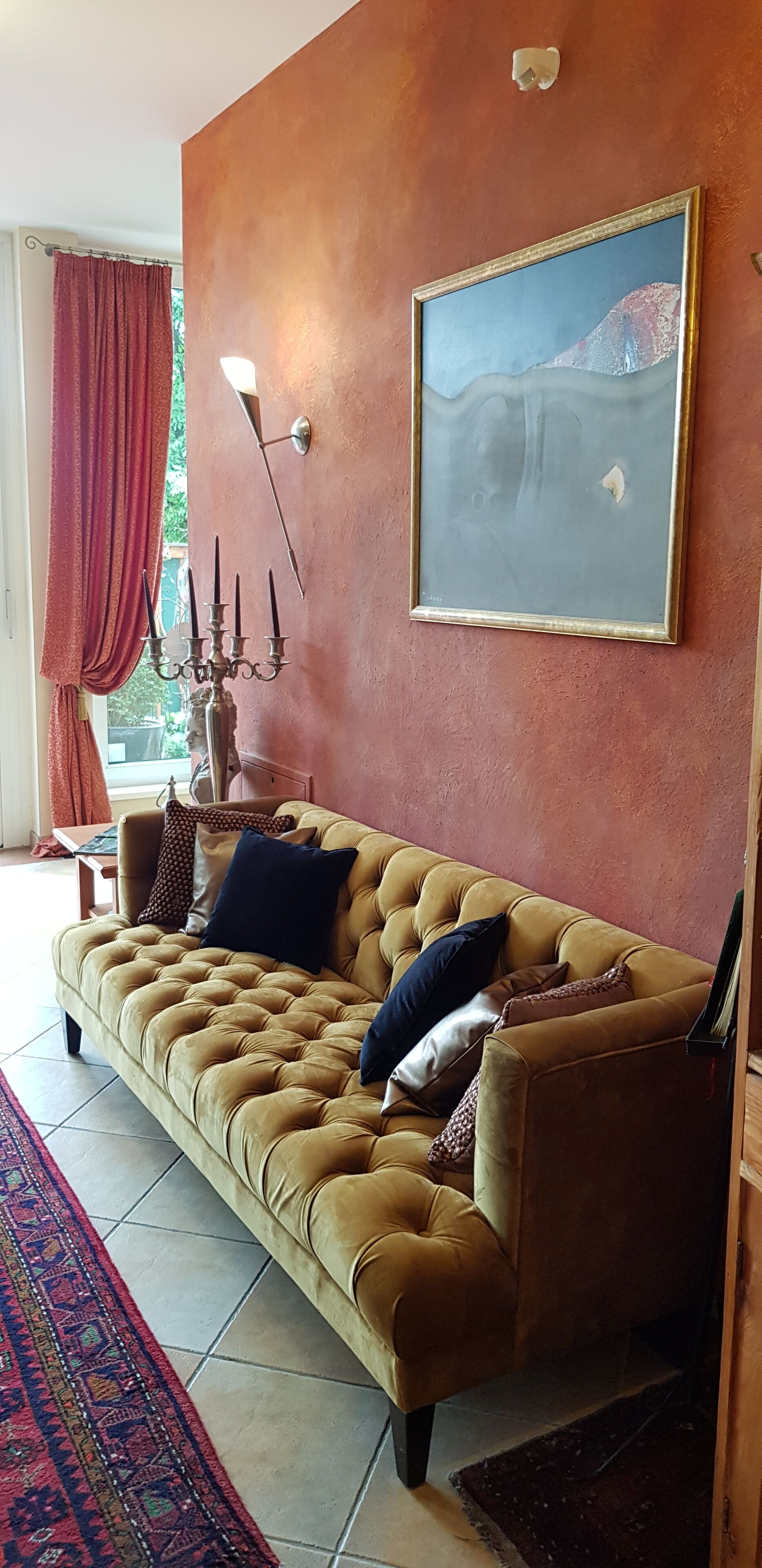 Un salón con un sofá beige, una alfombra estampada, un cuadro en la pared y una ventana con cortinas rojas.