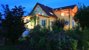Una casa de dos pisos con techo inclinado, varias ventanas y un jardín con diversas plantas y flores.