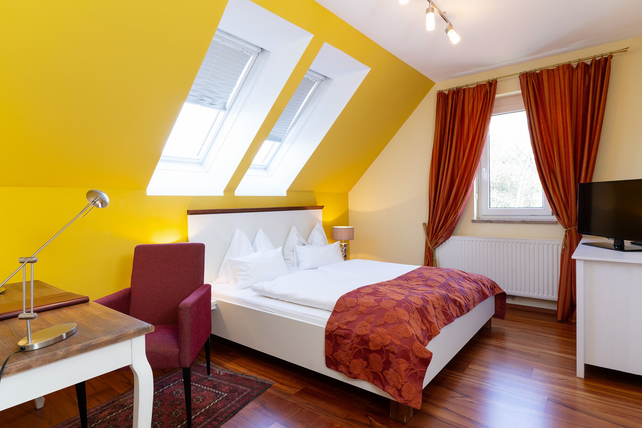 Ein Schlafzimmer mit Bett, Schreibtisch, Stuhl, Fernseher und einem Fenster mit Vorhängen.