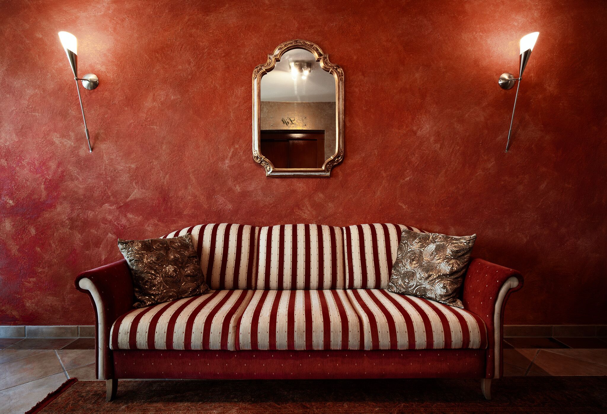 A red sofa with striped upholstery, a decorative mirror, and wall-mounted sconces against a textured wall.