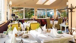 Comedor con mesas dispuestas para una comida, ventanas amplias con vistas a árboles y una araña.