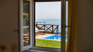 Vista a través de una ventana abierta de un área de piscina con terraza de madera y sillas de descanso.