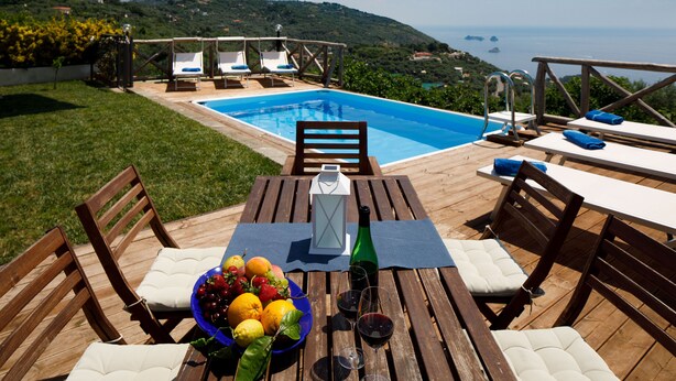 Una terraza de madera con piscina, mobiliario exterior y una mesa dispuesta con frutas y bebidas.