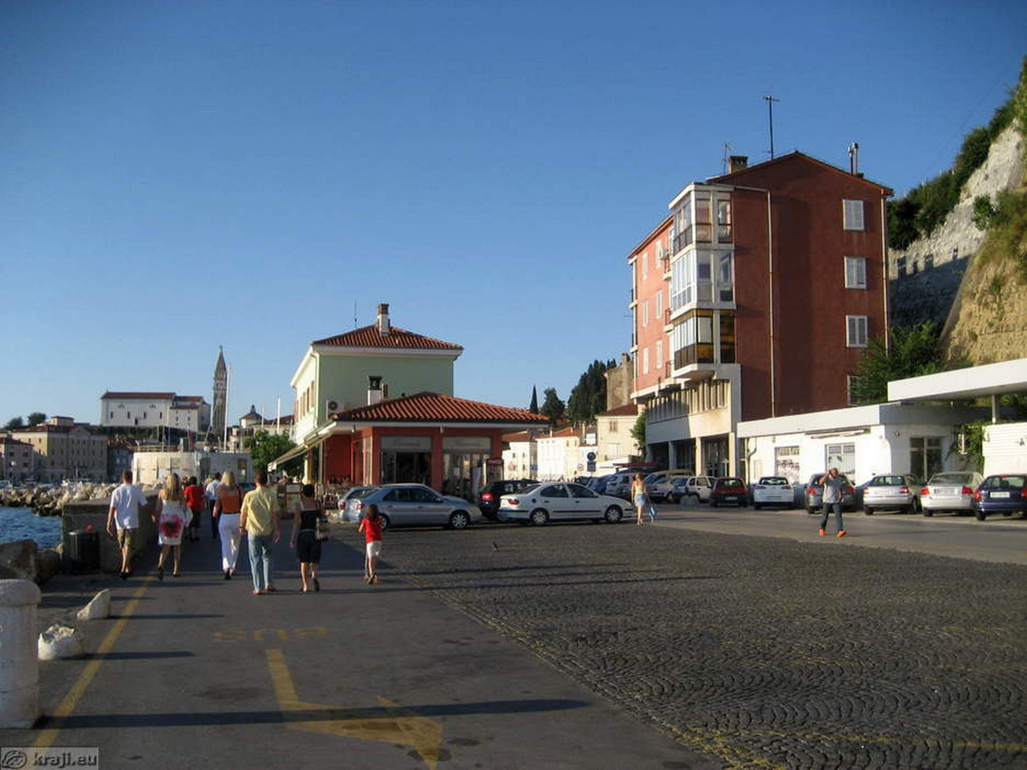 A coastal street with pedestrians, parked cars, and buildings including a prominent red brick building and a white building with a red roof.