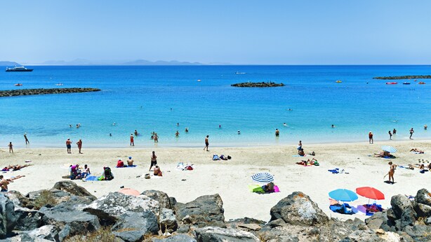 Une plage avec une eau d’un bleu limpide, un rivage rocheux, et des gens qui se prélassent au soleil et se baignent.