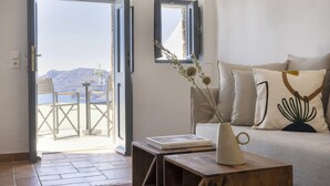 A room with a view of mountains through open doors, a sofa with decorative pillows, a vase with dried plants, and a book on a side table. - Bluedome Cavehouses by Otium Villas (Santorini)