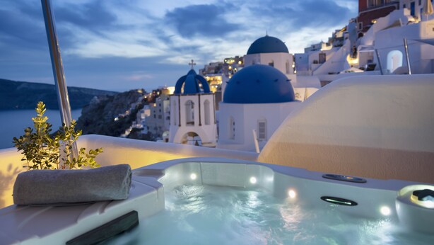 A hot tub with a towel and a plant, overlooking a coastal town with white buildings and blue domes. - Bluedome Cavehouses by Otium Villas (Santorini)