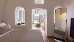 A bedroom with a large bed, a mirror, and a view of a dining area through an arched window. - Bluedome Cavehouses by Otium Villas (Santorini)