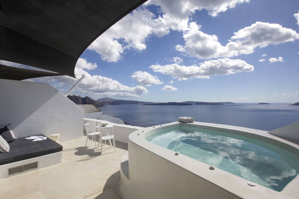 Un área de estar moderna al aire libre con jacuzzi, mobiliario blanco y vistas al mar y las montañas.