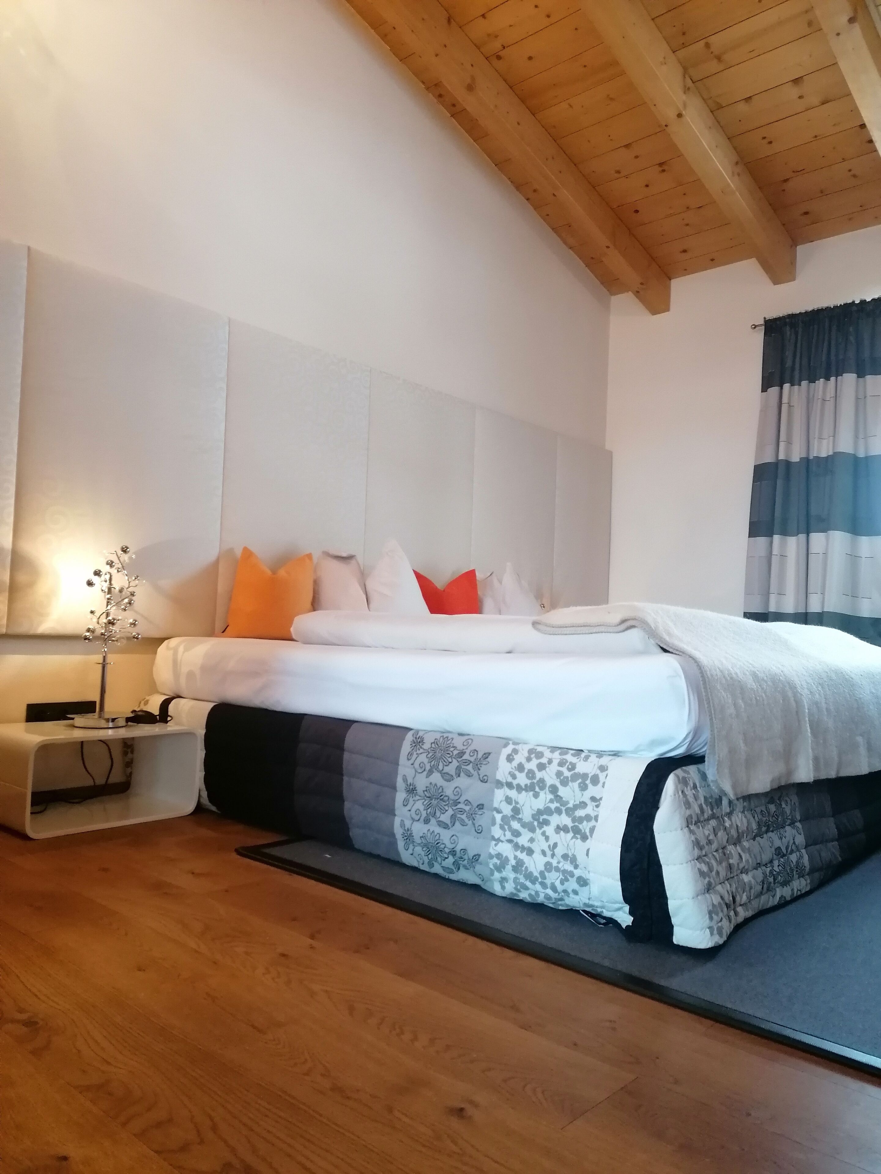 Een slaapkamer met een houten plafond, een bed met een wit hoofdeinde, een nachtkastje met een lamp en een raam met gestreepte gordijnen.