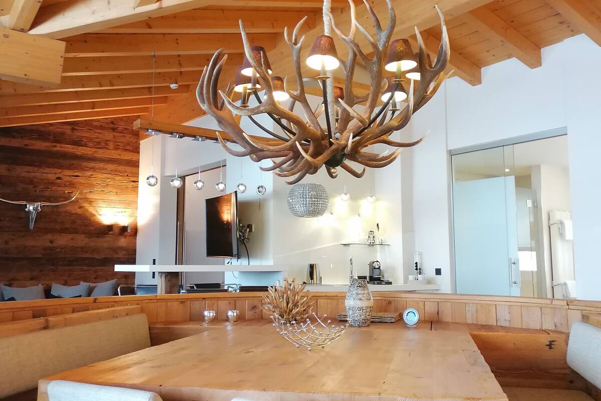Uma zona de refeições moderna com teto de madeira, um candelabro com braços semelhantes a galhos de veado e uma grande mesa de madeira com assentos.