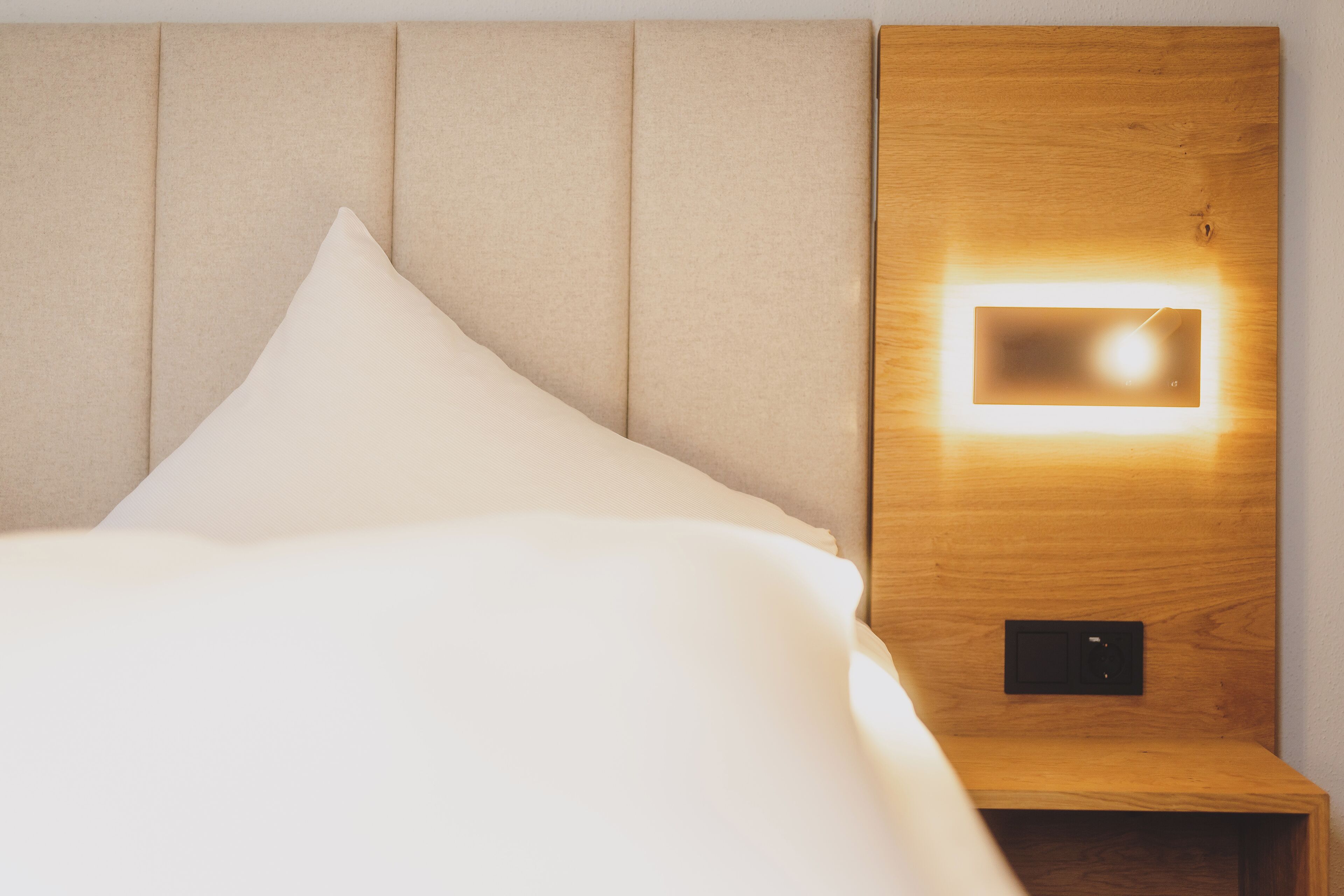 Un lit bien fait, avec une tête de lit, une table de chevet avec une lampe et une prise électrique.