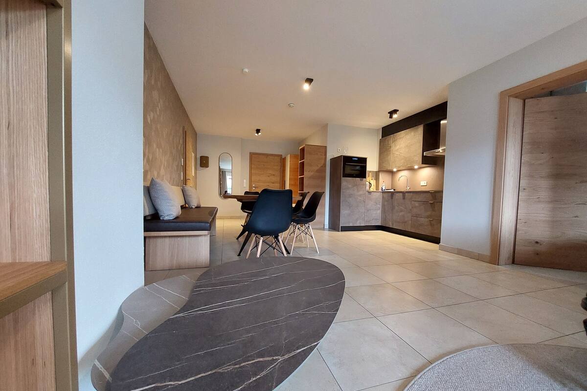Uma sala de estar moderna com um sofá preto curvo, uma área de jantar com mesa e cadeiras de madeira e uma cozinha com forno embutido e micro-ondas.