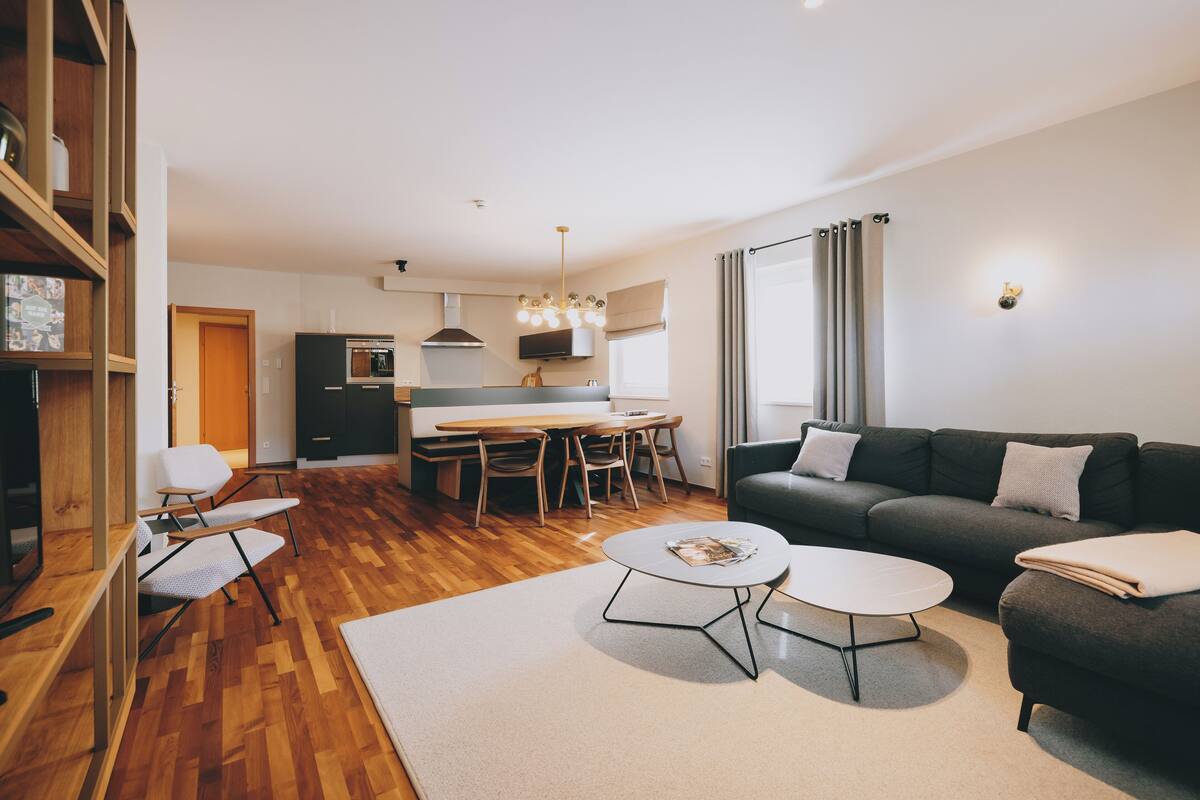Uma sala de estar moderna com sofá, mesa de centro e cadeiras. Há uma área de jantar com mesa e cadeiras. O ambiente possui piso de madeira e uma janela grande com cortinas.