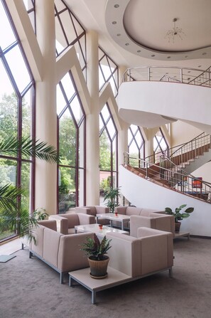 A spacious lobby with high ceilings, large windows, a central seating area, and a staircase leading to an upper level. - Park Hotel Odessos (Golden Sands)