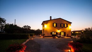 A two-story house with a gravel driveway, lit by warm outdoor lights, surrounded by potted plants and flowers. - Farmhouse in Maremma a few steps from the sea (Albinia)