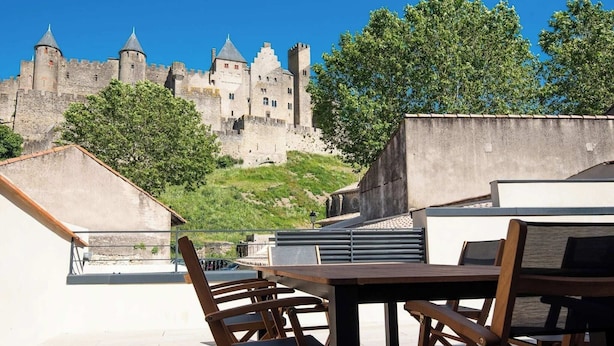 A rooftop terrace with a view of a historic castle, wooden furniture, and a clear sky.