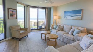 A spacious living room with a large sofa set, a small round table, and a chair. There is a balcony with a view of the ocean.