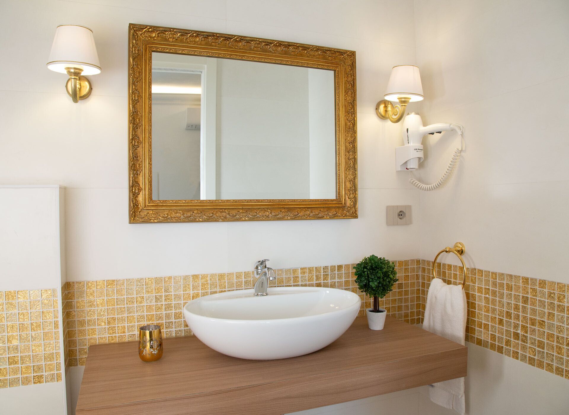 Un baño con un lavamanos ovalado grande, espejo con marco dorado y dos lámparas de pared.