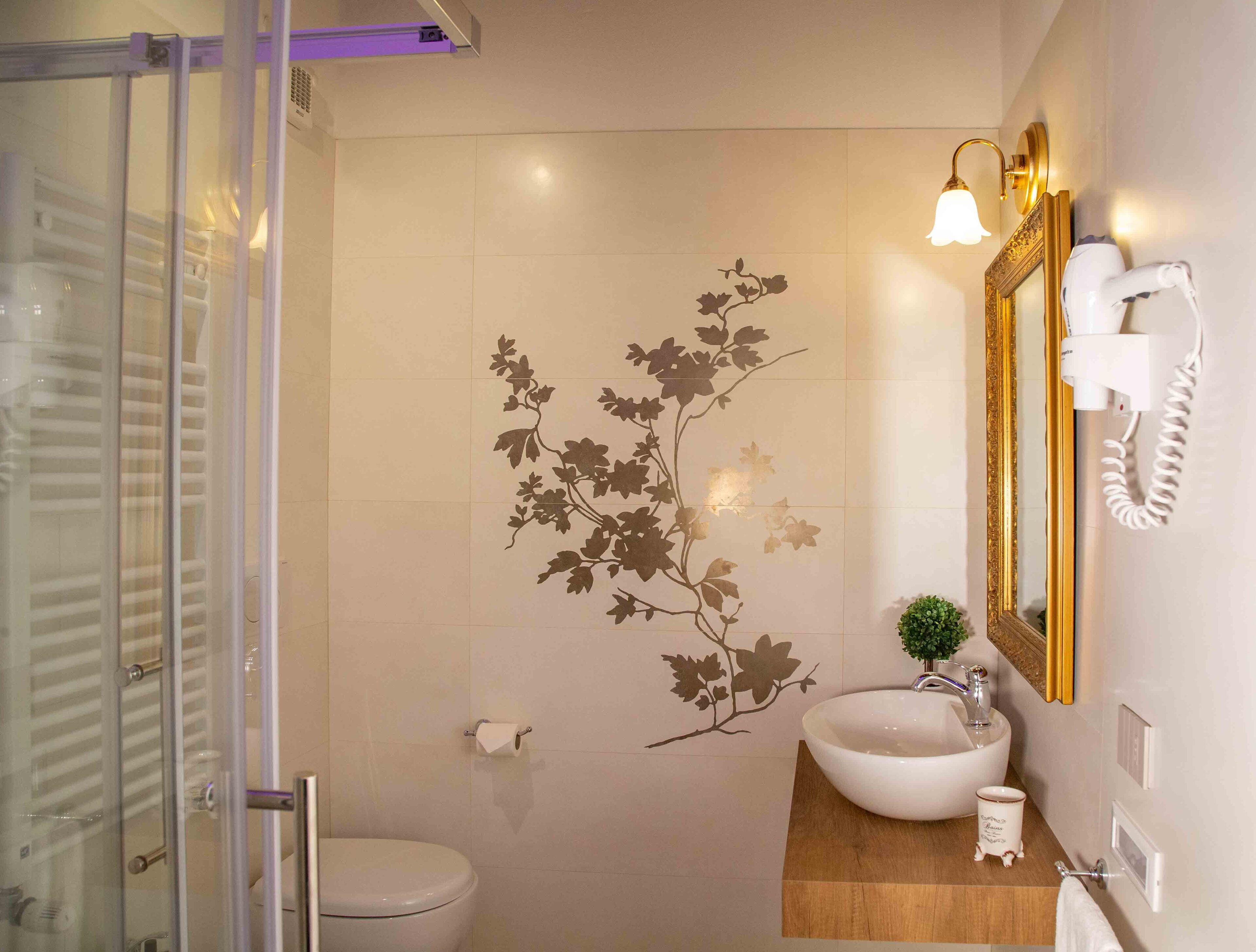 Un bagno con box doccia in vetro, un WC, un lavandino con bacino bianco, una piccola pianta e uno specchio con cornice dorata.