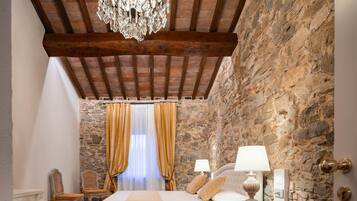 Un dormitorio con pared de piedra, vigas de madera, una araña, una cama con ropa blanca, dos mesitas de noche con lámparas y una ventana con cortinas.