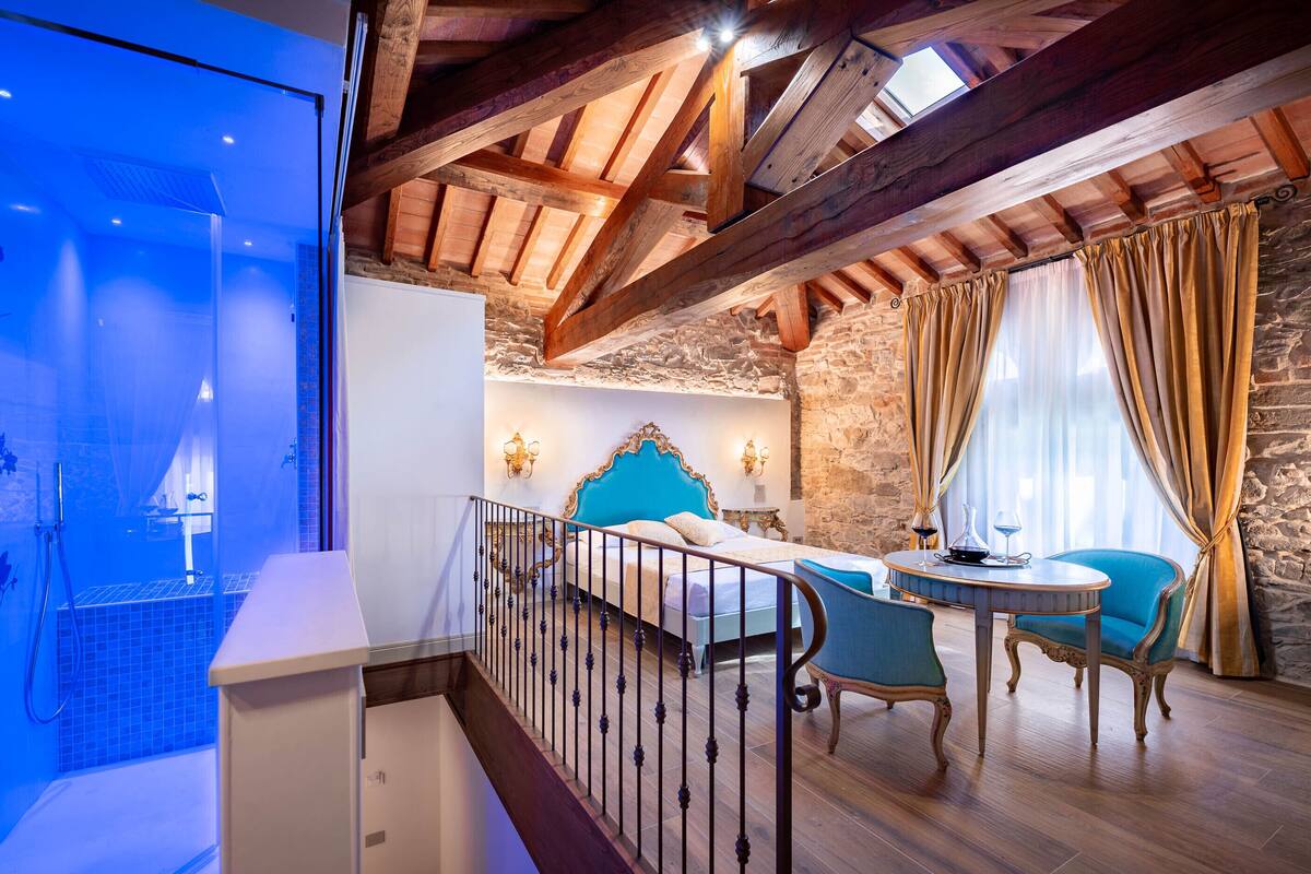 A bedroom with a large skylight, a bed with a blue headboard, a small table with two chairs, and a stone wall.