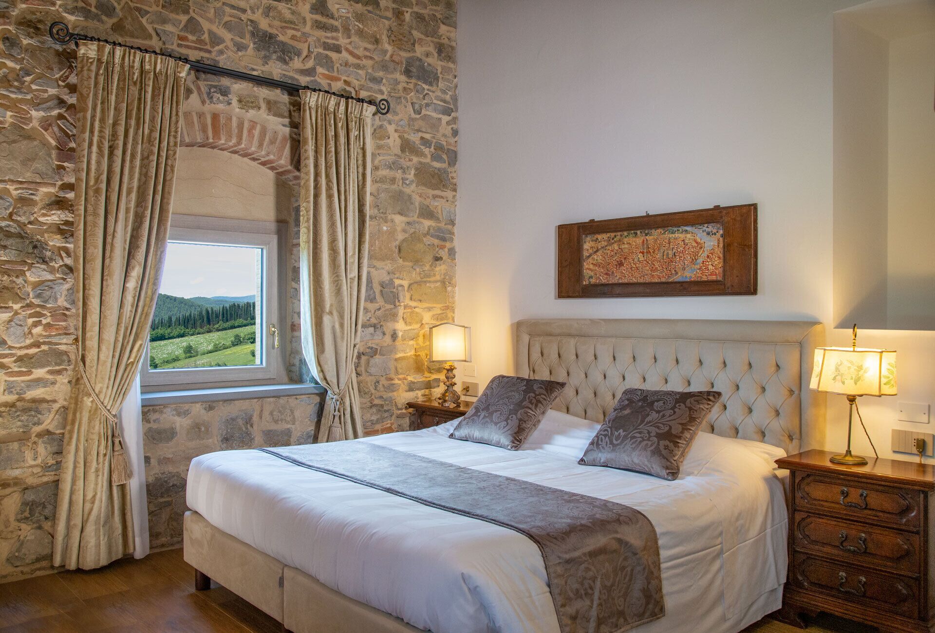 Un dormitorio con pared de piedra, una cama con cabecera, dos lámparas de noche y un ventanal con cortinas.
