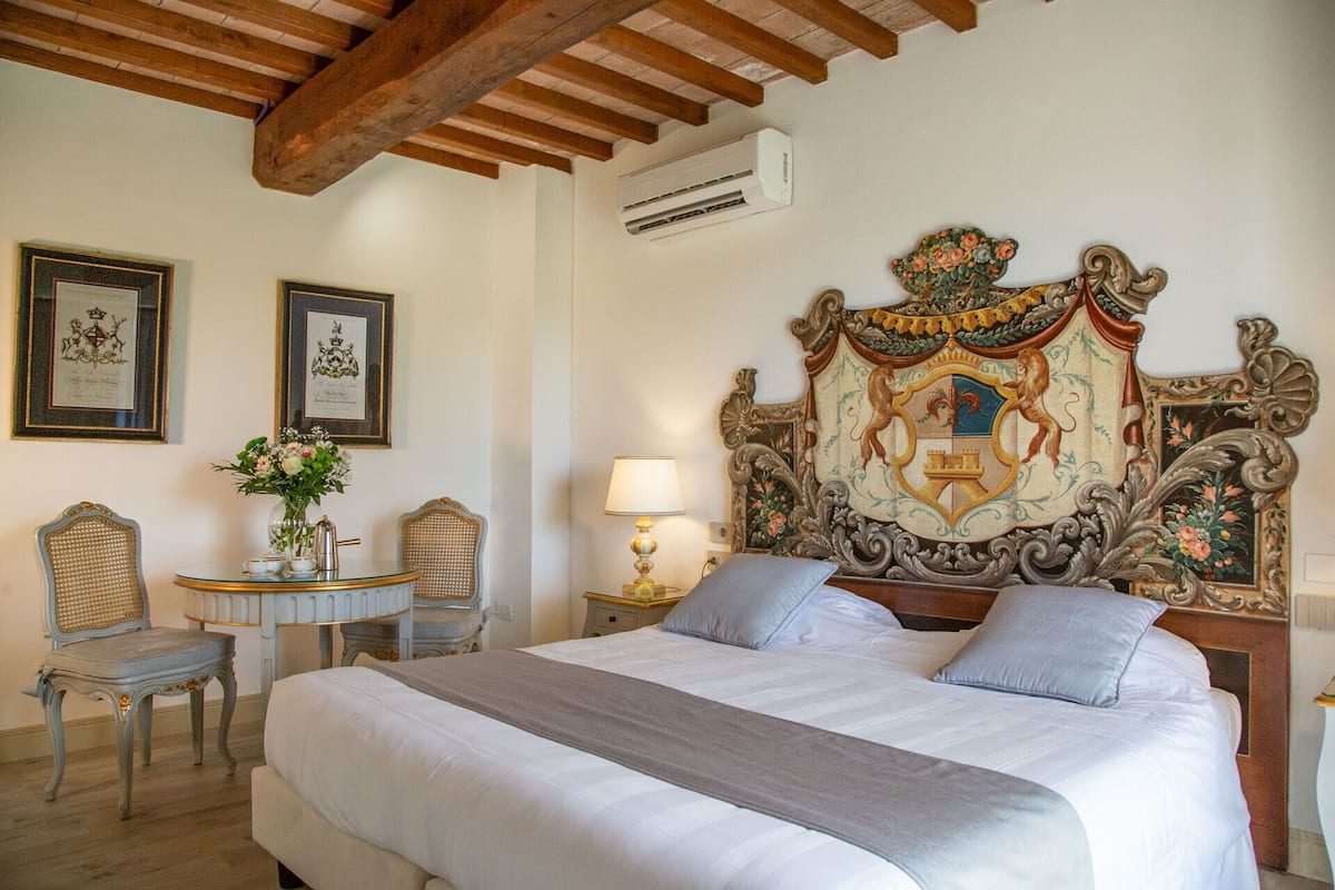 A bedroom with a large bed, two chairs, a small table, and ornate headboard.