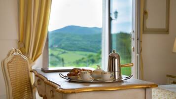 A breakfast setup with a tray of croissants, cups, and a coffee pot on a table by a window with a view of green hills.