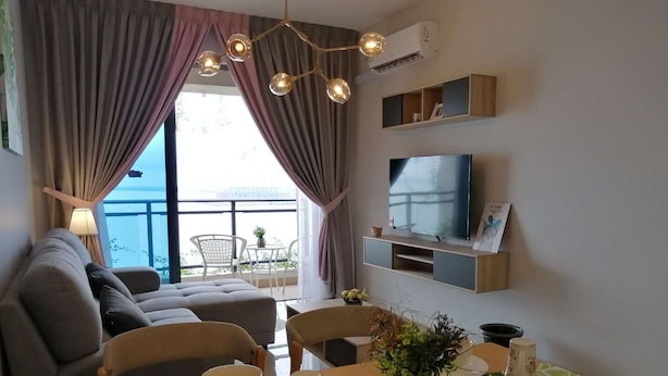 A modern living room with a sofa, dining table, and chairs. There is a TV mounted on the wall and a balcony with a view. - Forest City Ataraxia Park 4  by Wastone (Gelang Patah)