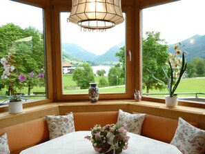 A room with a view of mountains and a lake, a table with a vase of flowers, and a bench with floral cushions. - Vacation Apartment in Weitl am See, with a picture window - Ferienwohnungen Weitl am See (Schliersee)
