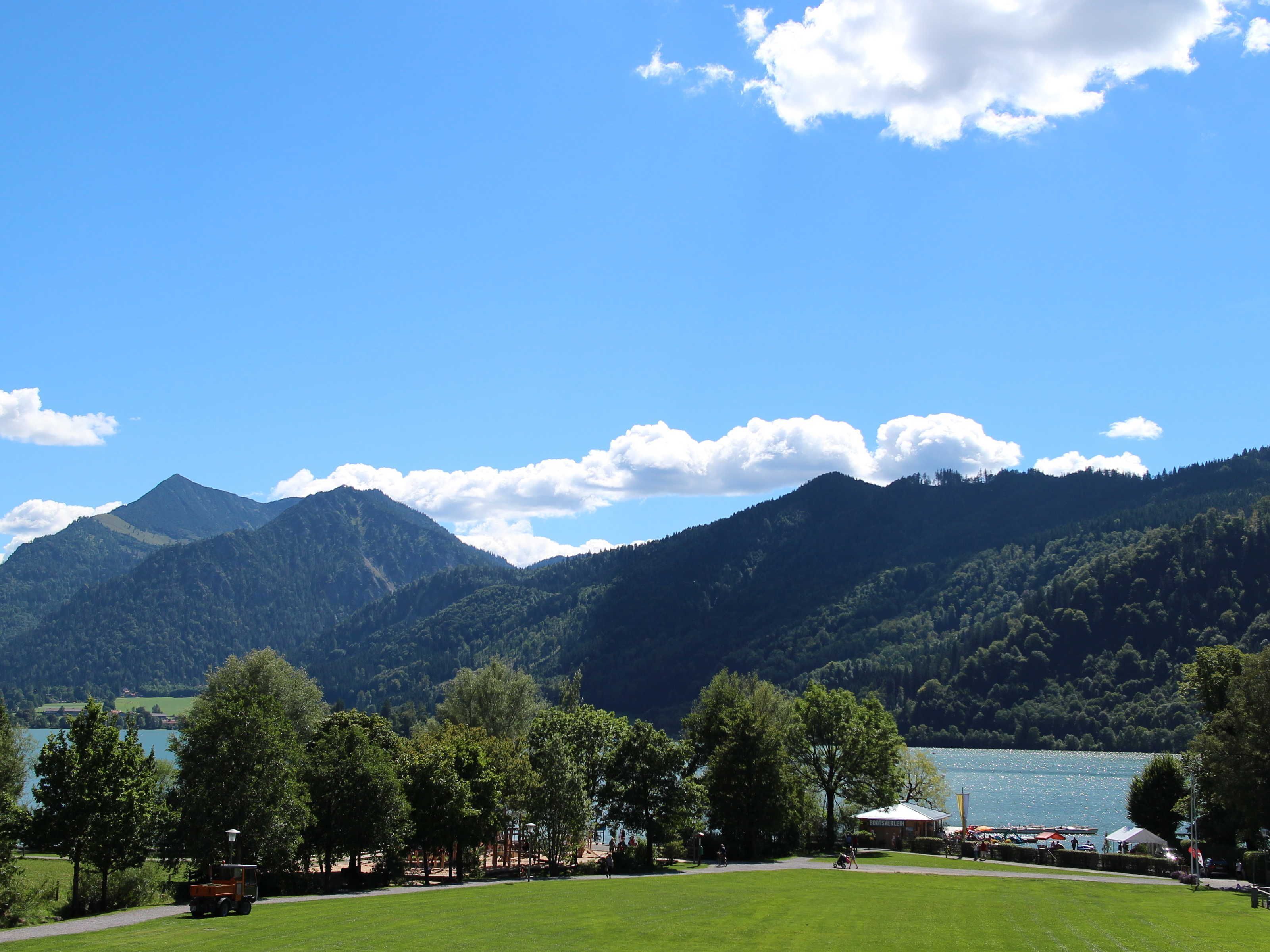 Una vista panoràmica d'un llac envoltat de muntanyes, amb una zona verda i alguns arbres al primer pla.