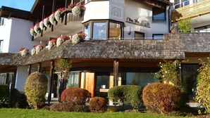Un immeuble à plusieurs étages, doté de balcons ornés de fleurs, d’une enseigne indiquant qu’il s’agit d’un hôtel et d’un jardin bien entretenu.