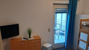 Una habitación pequeña con un armario de madera, un televisor, una mesita con una planta, una silla blanca y vistas a un balcón exterior con sillas.