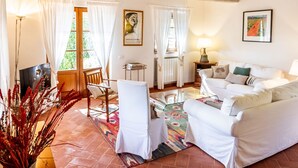Ein gemütliches Wohnzimmer mit einem weißen Sofa, einem Holzstuhl, einem Glastisch und einem Fernseher. Ein Fenster mit Vorhängen, eine Heizung und eine Lampe auf einem Beistelltisch sind ebenfalls vorhanden.