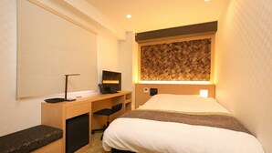 A compact hotel room with a bed, a desk with a computer, a chair, and a window with blinds.