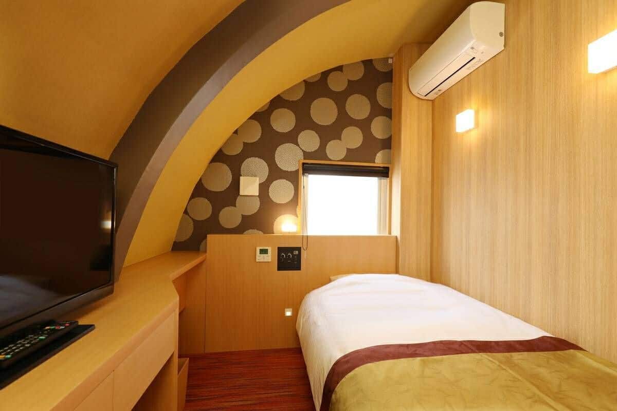 A compact hotel room with a bed, a television, an air conditioning unit, and a window with a view.