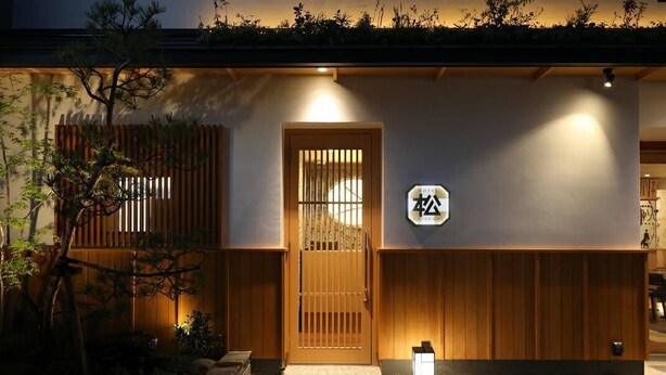 A traditional Japanese-style building with a wooden door, a sign with Japanese characters, and a small lantern on the ground.