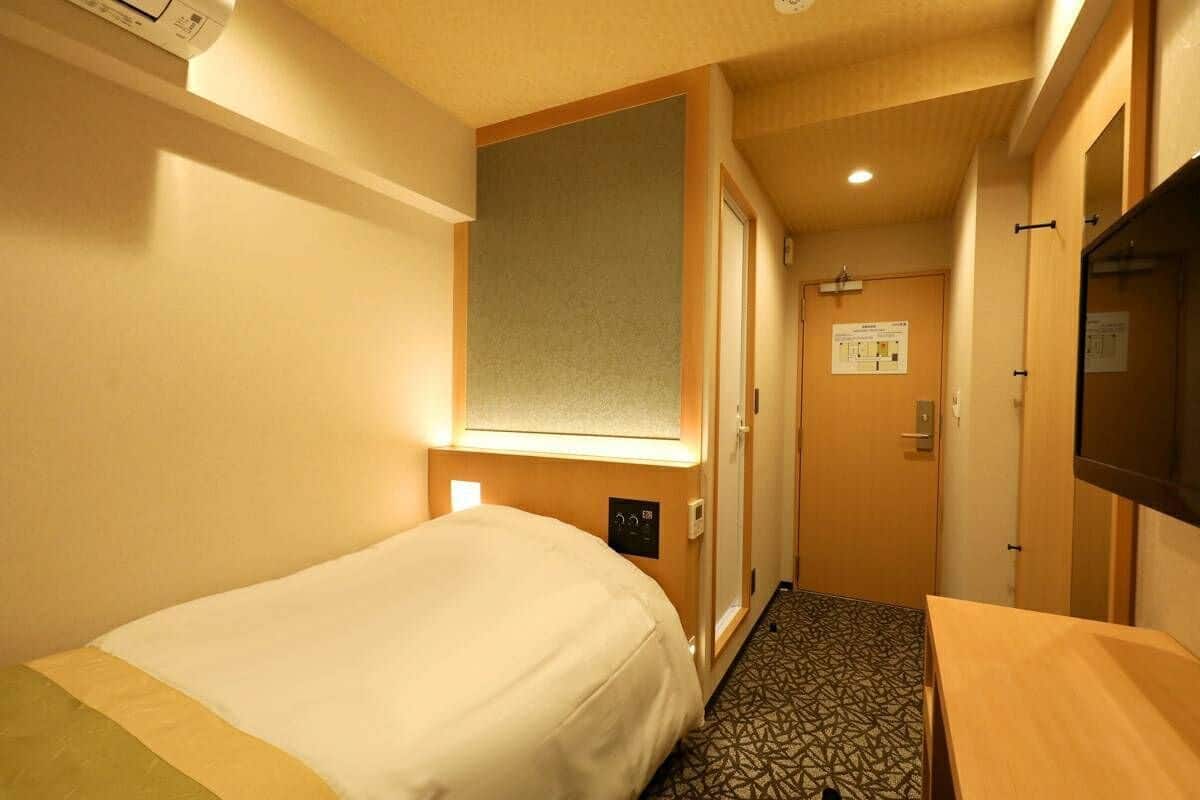 A hotel room with a bed, a small table, a wall-mounted TV, a door with a sign, and a patterned floor.