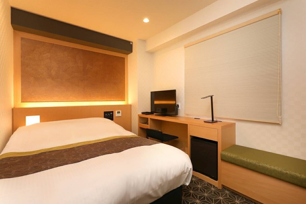 A hotel room with a bed, a desk with a computer, a bench, and a window with blinds.