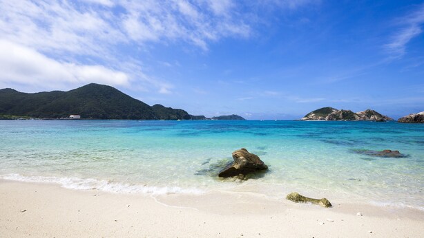 Een strand met helder turquoise water, een zandstrand en rotsformaties in de verte.