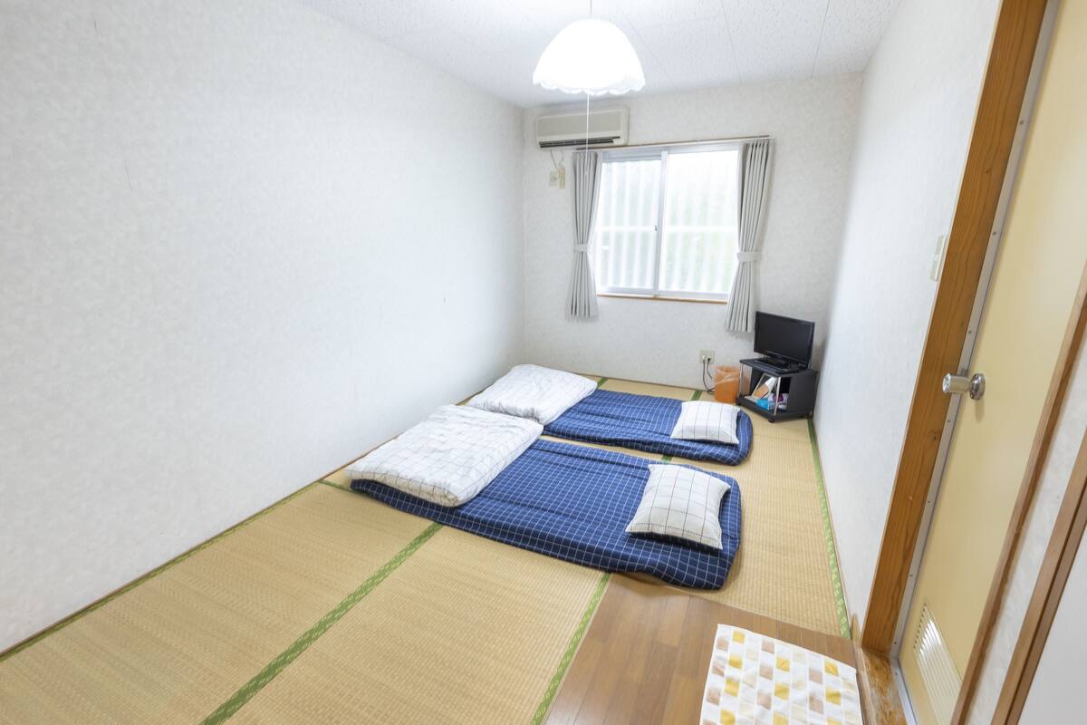A small, clean room with two beds, a window with curtains, a desk with a computer, and a door.