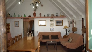 A cozy living room with a wooden dining table, a sofa, and a small table. There are shelves with various items and a ceiling light fixture. - Salvador's barrack (Canyadora)