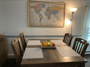 Une table à manger avec un plateau de fruits, des chaises en bois, une carte du monde accrochée au mur et un lampadaire.