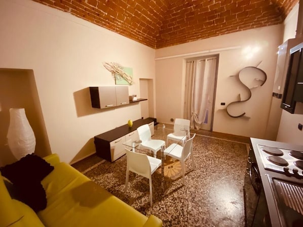 Un espacio habitable compacto con área de cocina, una mesa de comedor con sillas, un sofá amarillo y techo de ladrillo.