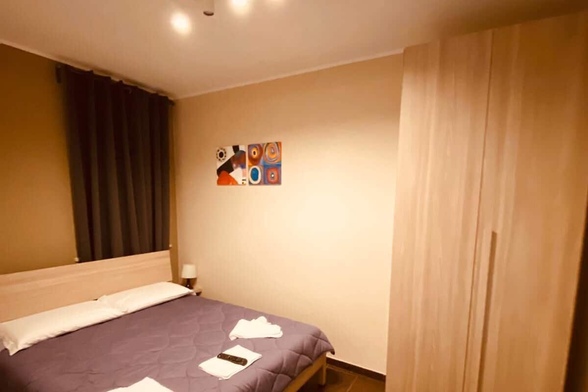 Ein ordentlich bezogenes Bett mit einer violetten Tagesdecke, weißen Kissen und gefalteten Handtüchern. Rechts daneben befindet sich ein Holzschrank und an der Wand ein farbenfrohes abstraktes Gemälde.