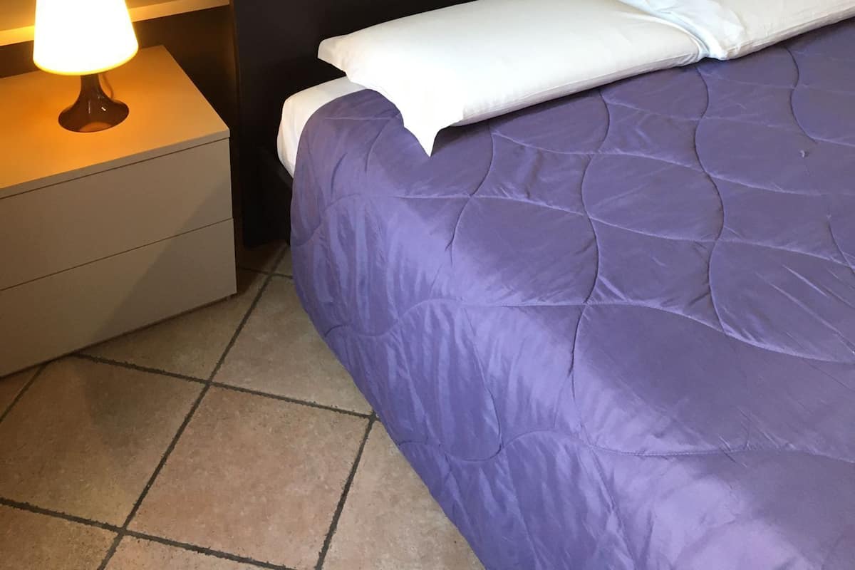 Ein ordentlich bezogenes Bett mit dunklem Kopfteil, weißen Kissen und einer violetten Decke. Es gibt zwei Nachttische mit Lampen, einen auf jeder Seite des Bettes.