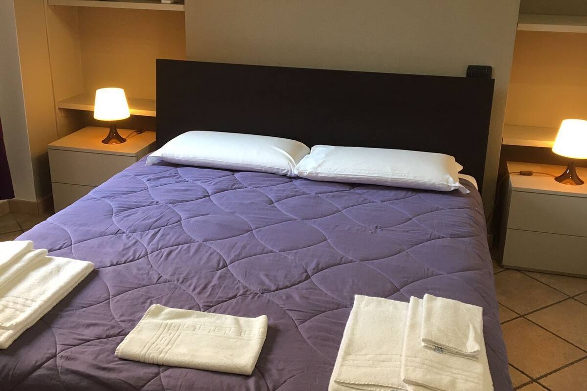Ein ordentlich bezogenes Bett mit einer violetten Steude, weißen Kissen und gefalteten weißen Handtüchern. An beiden Seiten des Bettes stehen je eine Nachttischlampe.