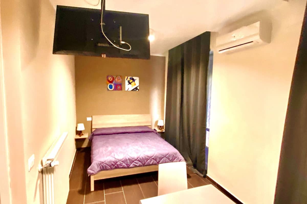 Ein kleines, schmales Hotelzimmer mit einem Bett, einem kleinen Tisch, einem an der Wand montierten Fernseher und einem Heizkörper.