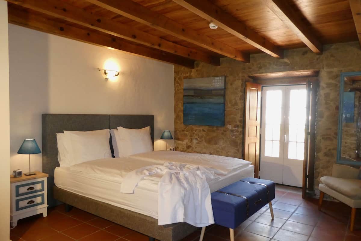 A bedroom with a large bed, wooden ceiling beams, stone walls, a nightstand with a lamp, and a bench.